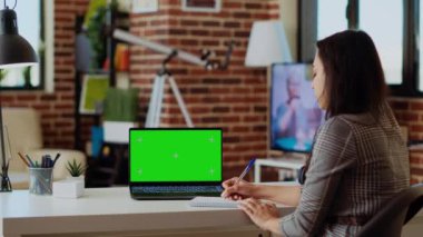 Evde kalan tele-işçi, ahşap masadaki çeşitli işleri yapmak için yeşil ekran laptopunu kullanıyor. Krom tuşlu klavyeye veri yazıp not defterine yazan uzak çalışan bir çalışan