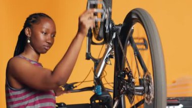 Afrikalı Amerikalı kadın pedal çevirerek bisiklet performansını kontrol ediyor. Turuncu stüdyo arka planında bisiklet tekerleklerinin düzgün çalışmasını sağlayan profesyonel kamera A