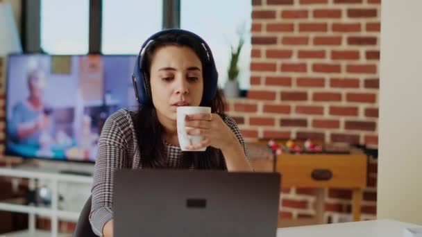 インド人女性がコーヒーを飲んで ヘッドフォンを使って自宅でテレビ番組を見ている ラップトップ カメラBでテレビシリーズを楽しんでいる間 熱い飲み物で目覚めている人 — ストック動画