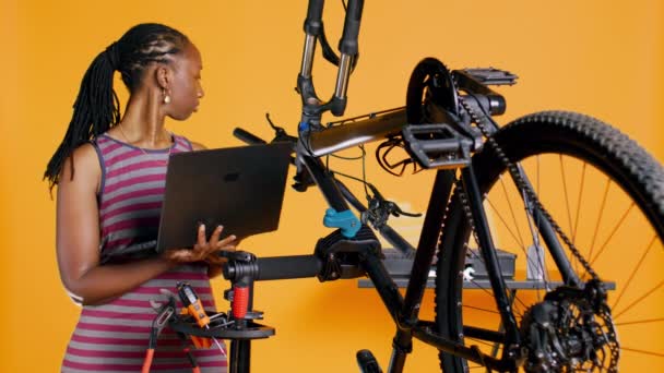 アフリカ系アメリカ人エンジニア スタジオのバックグラウンド修理工場で自転車サービスを行い ノートパソコンのメンテナンスリストを見ています 修理を必要とする専門の点検自転車の部品 カメラB — ストック動画
