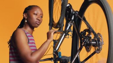 BIPOC mühendisi pedalları çevirerek bisiklet performansını kontrol ediyor. Turuncu stüdyo arka planında bisiklet tekerlerinin düzgün çalışmasını sağlayan uzman, kamera A