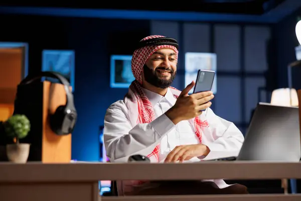 彼のデジタルモバイルデバイス上でネットをサーフィンする熱狂的なイスラム男性 スマートフォンを使用してテーブルに座って笑顔で男性イスラム教徒の詳細なビュー — ストック写真
