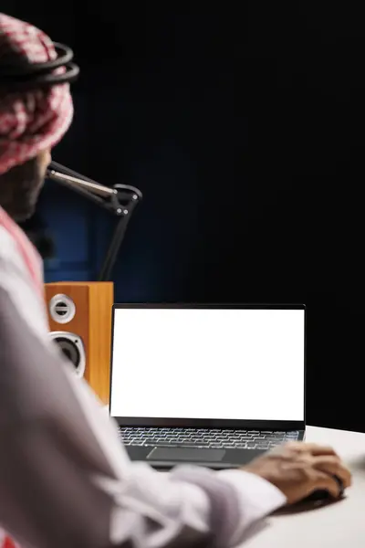 画像は 自宅で働くイスラム教徒の男性が 自分の机に座って ミニコンピュータに示された単離染色体のテンプレートを描写しています コピースペースの白いスクリーンが付いているラップトップを使用して若いアラブの男 — ストック写真