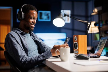 Oturma odasında, Afrikalı Amerikalı adam Webinar dersini izlerken cep telefonu uygulamasından mesaj gönderiyor. Bir yönetim kursu kapsamında muhasebe ve mali konuları okuyan genç kişi.