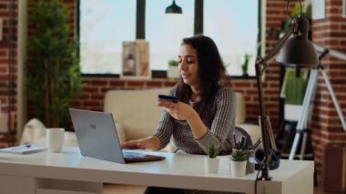 Gülümseyen kadın online alışveriş yapmaktan zevk alıyor, dizüstü bilgisayarına bankamatik kartı bilgilerini dikkatlice yazıyor. Web sitesine ödeme yöntemi eklenirken, modern apartman oturma odasında, kamera B