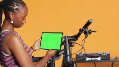 Model tabletle özel ders videolarını izleyen neşeli kadın bozuk bisikleti tamir etmeyi öğreniyor. Gülümseyen bisiklet hobisi bisikleti tamir ederken eğleniyor, yeşil ekran cihazında online rehber takip ediyor, kamera A