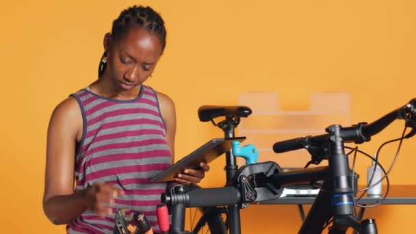 タブレット上のインターネットチュートリアルに続く陽気な女性は 彼女の壊れた自転車を修復することを学びます 楽しい自転車のコンポーネントを修理する楽しい自転車の趣味を持つハッピーサイクリング趣味 オンラインガイドに従って スタジオの背景 — ストック動画