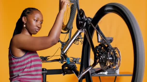 アフリカ系アメリカ人女性がペダルを回転させて自転車の性能をチェック オレンジのスタジオの背景 カメラBの自転車の車輪のタイヤの適切な機能を保障する専門家 — ストック動画