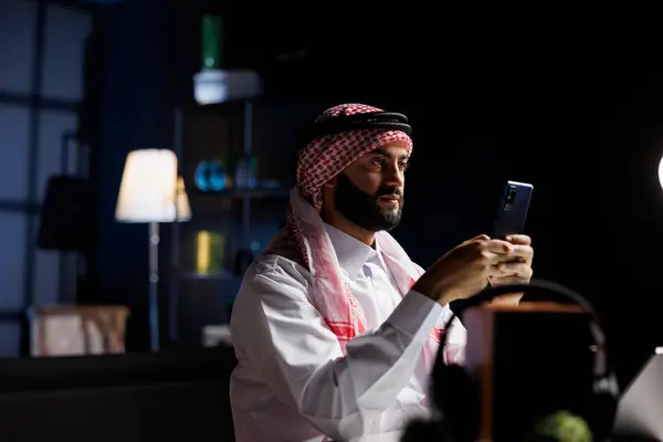 中東の起業家は オンラインコミュニケーションと研究のためにスマートフォンを使用して 彼のオフィスデスクで勤勉に働いています 画像は デジタルモバイルデバイスを保持しているテーブルにアラブの男を示しています — ストック写真