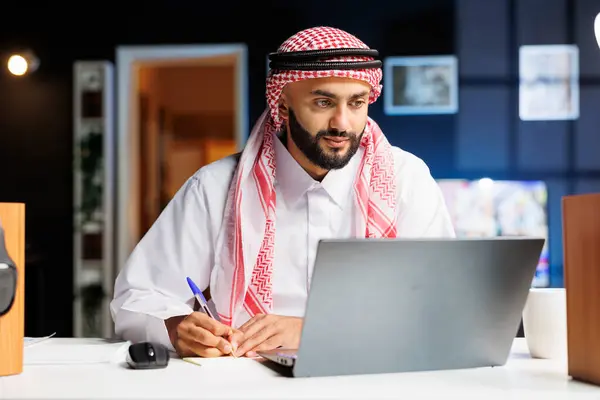 アラブの起業家は よく組織されたオフィスデスクで自分のラップトップに効率的に取り組んでいます 彼はタイプし インターネットを閲覧し 現代の技術に囲まれてノートを取ります — ストック写真