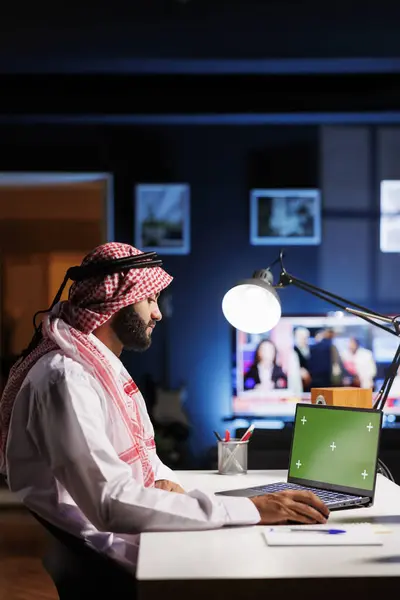 伝統的な服装を身に着けているアラブの男性プロが 空の緑色のスクリーンを備えたラップトップを使用して オフィスデスクに座っています 画像は 広告やプレゼンテーションのための現代的なワークスペースを描いています — ストック写真