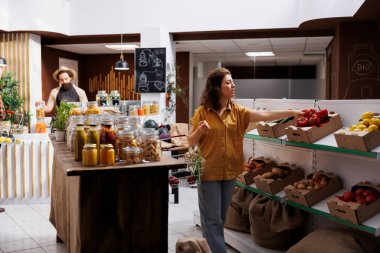 Green living woman in zero waste store interested in purchasing natural vegetables with high nutritional value. Client does grocery shopping in sustainable local neighborhood supermarket clipart