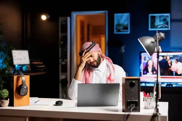 オフィスデスクに座っているアラビアの衣料品のフラットビジネスマンは オンライン研究のためにラップトップを使用しています テーブルの上のミニコンピュータを持つ若いイスラム教徒の男は頭痛で疲れているように見えます — ストック写真