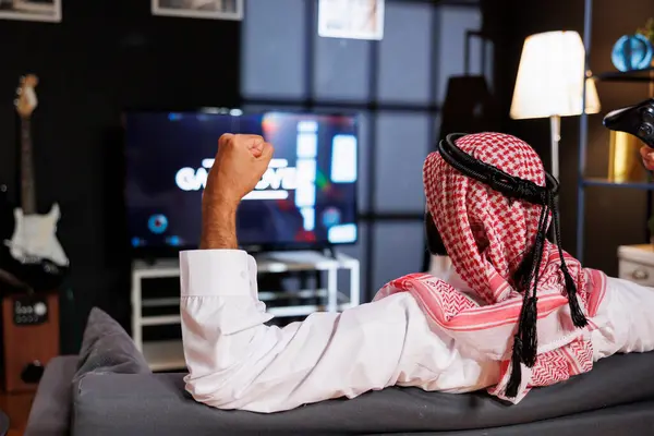 快適なソファーに座っている間 空中に拳で勝利を祝うアラブ男性の詳細なイメージ ワイヤレスコントローラを把握した中東男性のリアビューショット — ストック写真