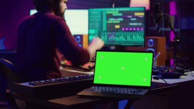 Müzik mühendisi konsol ve daw yazılımını karıştırma üzerine ses kayıtlarını düzenliyor, ev stüdyosunda yeşil ekranlı bir laptop var. Sanatçı, akustik mühendislik araçlarıyla yeni bir şarkı üretiyor. Kamera A.