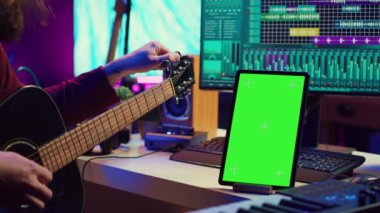 Müzik yapımcısı çalmadan önce gitarını akort etmeyi öğreniyor, tablette yeşil ekran ekranı kullanarak ders videosu izliyor. Genç sanatçı telleri akort ediyor, pratik yapıyor. Kamera A.