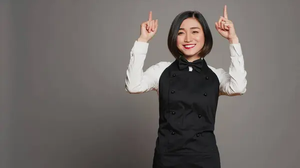 Asian woman pointing at something upwards in studio, indicating direction above her head. Waitress server with apron showing up indication over grey background, table service. Camera A.
