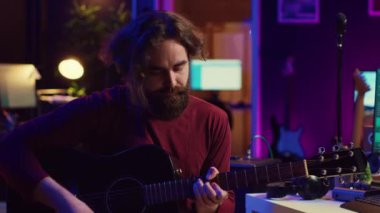 Genç adam gitar çalıyor ve daw yazılım arayüzünü kullanarak müzik yapıyor. Müzik yapımcısı stüdyoda şarkı kaydediyor ve stereo konsolda akustik sesleri karıştırıyor. Kamera A.