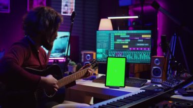 Erkek sanatçı online ders programında gitar çalmayı öğreniyor, akustik enstrüman çalışıyor ve yeşil ekran ekranlı telefona bakıyor. Müzik yapımcısı gitar çalacak. Kamera B.