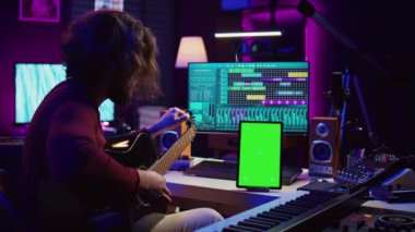 Müzik yapımcısı çalmadan önce gitarını akort etmeyi öğreniyor, tablette yeşil ekran ekranı kullanarak ders videosu izliyor. Genç sanatçı telleri akort ediyor, pratik yapıyor. Kamera B.