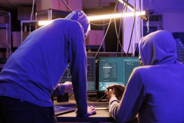 Güvenli verileri kırmak için bilgisayar ekranında kötü niyetli yazılım çalıştıran iki hacker. Suç örgütü kanunları çiğnerken kapüşon takıyor ve terk edilmiş depoda birlikte çalışıyorlar.