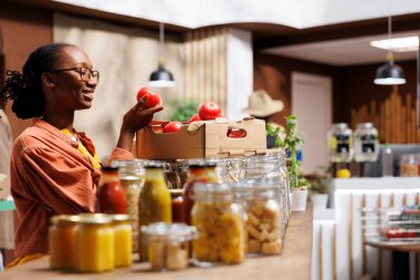 Photo focus on the smiling black woman in a bio-food store examining an organic farm-grown tomato. Client of African American ethnicity joyfully admires fresh produce in the eco friendly market. clipart