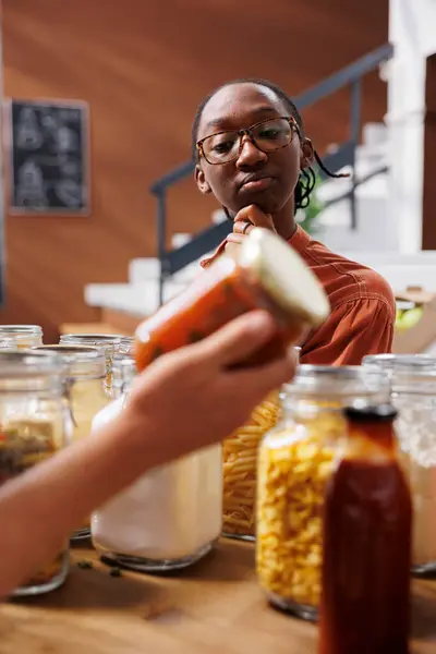 African American customer admires glass jar filled with organic red liquid food sauce that someone else holds in front of her. Black woman thinking of different recipes to make with bio food product.