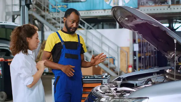 検査後 車内で見つかったクライアントの損害を示す認可された自動修理工場の従業員 アフリカ系アメリカ人のプロフェッショナルが彼女の車に含まれる油漏れについて女性に話す — ストック写真