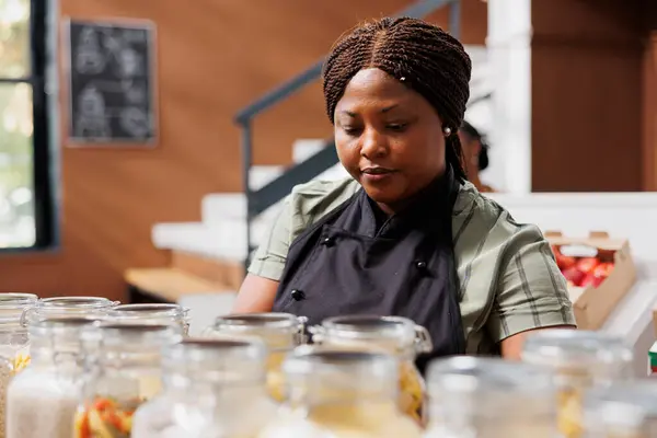 Portrait of black woman wearing an apron working in eco friendly grocery store, arranging organic sustainable items. African american seller organizing glass jars filled with healthy preserved items.