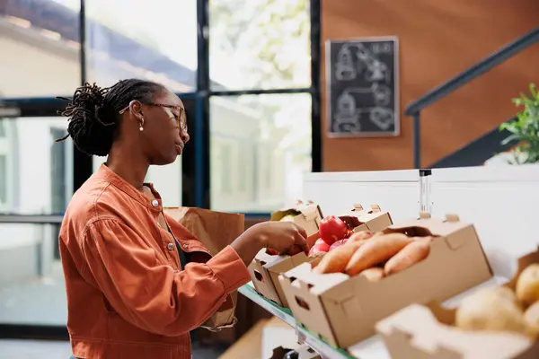 戴眼镜的黑人妇女从环保商店货架上的箱子里挑选当地生产的农产品 研究不同新鲜水果和蔬菜的非裔美国女性个体 — 图库照片