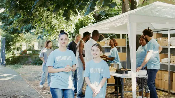 stock image Portrait of a woman and daughter taking part in food bank program to fight hunger. Blue-shirted Caucasian volunteers are ready to help less fortunate while glancing into camera. Handheld shot.