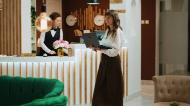 Lobi salonundaki dizüstü bilgisayarını kullanan üst düzey bir kadın web sitesindeki ünlü simgelere bakmak için internet ağında geziniyor. Otel misafiri otele gelir ve odayı görmek için bekler. El kamerasıyla..