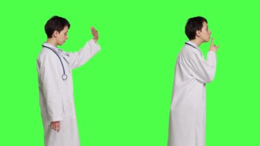 Genel pratisyen, sessiz sessizlik işareti gösteriyor ve insanları yakına gelmeye çağırıyor. Beyaz önlüklü, yeşil perde arkaplanına karşı duruyor. Doktor sessiz olmak istiyor, birini davet ediyor. Kamera B.