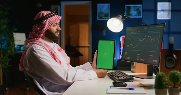 stock image Muslim worker in apartment office holds chroma key tablet, crosschecking data on it with business report graphs on computer. Smiling entrepreneur with mockup digital device in hand performs job tasks