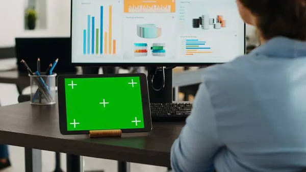 Company employee having tablet on desk with greenscreen display in startup agency office, using blank mockup template and isolated chromakey layout. Woman looking at digital web app on device.