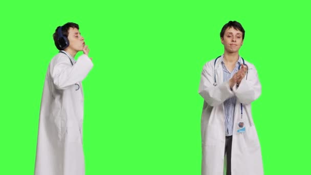 全科医生拍手 在耳机上听音乐 站在绿屏的背景下 女专家为某人喝彩 用清凉的歌声跳舞 摄像头B — 图库视频影像