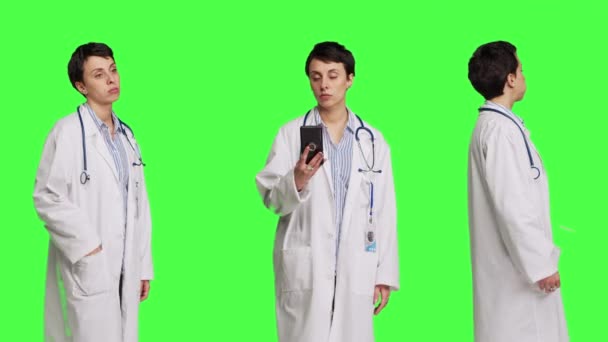 女性の専門家が携帯電話の時計を見て 患者が試験の予定に到着するのを待っています スタジオで緑色の背景 白いコートの一般的な実践者は 不快に動作する カメラ — ストック動画
