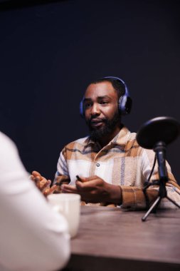 Siyahi erkek sunucu, online talk şovu için modern mikrofon ve kablosuz kulaklık kullanan biriyle röportaj yapıyor. Afrikalı Amerikalı bir adam podcast bölümü kaydeden biriyle konuşuyor..