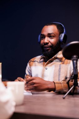 Afrikalı Amerikalı adam evde online radyo programı yapmak için ses ekipmanı kullanıyor. Kablosuz kulaklıklı ve mikrofona konuşan genç siyahi erkek içerik yaratıcısı bir podcast yapıyor..