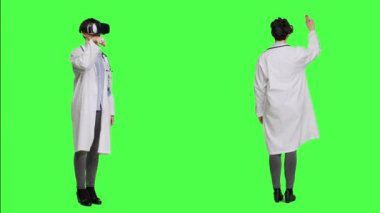Kadın sıhhiyeci stüdyoda üç boyutlu sanal gerçeklik kulaklığıyla çalışıyor, yeşil ekran arka planına karşı duruyor. Pratisyen hekim, VR gözlüklerini etkileşimli görme aracı ve fütüristik muayeneyle kullanır. Kamera A.
