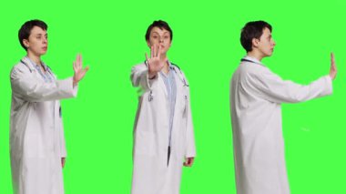 Stüdyoda el kaldıran ve dur işareti yapan kadın doktor, yeşil perde arkaplanına karşı ret ve inkarı ifade ediyor. Pratisyen hekim uyarı sembolünü olumsuz tepkiyle gösteriyor. Kamera B.