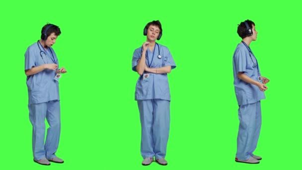 陽気なヘルススペシャリストは オーディオヘッドセットでクールな音楽を聴き 緑色の背景と踊り 楽しんでいます 楽しいダンスの動きをする歌を楽しむ看護師 レジャー カメラ — ストック動画