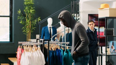 Afrika kökenli Amerikalı soyguncu şık kıyafetler çalmaya çalışıyor, güvenlik copu ile tehdit ediliyor ve modern butikte Asyalı koruma satın alıyor. Güneş gözlüğü ve başlık takan hırsız tanınmayacak halde.