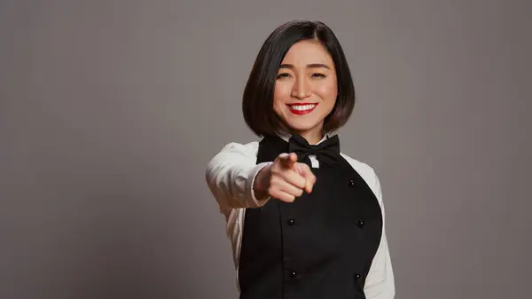 Asian waitress pointing at camera to choose you over grey background, wearing uniform with bow and apron. Woman catering worker choosing someone to work at a restaurant. Camera B.