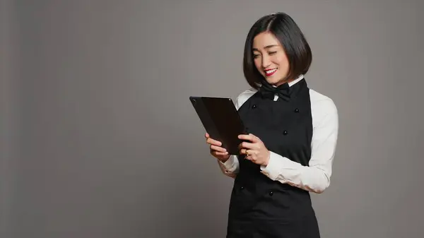 Asian restaurant server checking all table reservations on tablet, browsing through clients information on website. Waitress looking at online webpages to see availability. Camera A.