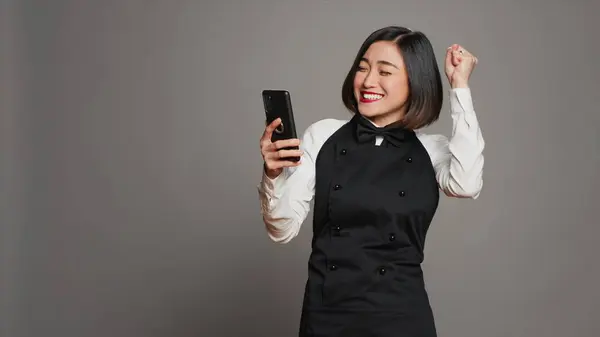 Asian server feeling happy after reading good news on phone, having a lot of clients with reservations for dinner. Waitress with apron using smartphone to see table bookings, enjoys work. Camera A.