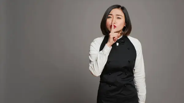 Restaurant hostess showing silence symbol in studio, doing hush mute gesture with finger over lips. Private waitress asking to keep secret with confidential information, secrecy sign. Camera A.