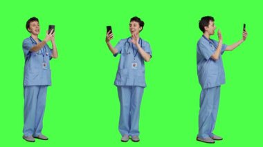 Hemşire iş molasında video görüşme bağlantısına cevap veriyor, akıllı telefonu kullanarak arkadaşlarıyla çevrimiçi video konferans sohbetinde bağlantı kuruyor. Önlük uzmanı yeşil ekrana karşı duruyor. Kamera A.