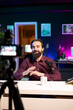 Neşeli internet yıldızı profesyonel kamera önünde konuşuyor ve stüdyoda kaliteli mikrofon, izleyicilerle tartışıyor. Neşeli adam sosyal medya videolarını kaydetmek için yüksek teknoloji kayıt cihazları kullanıyor.