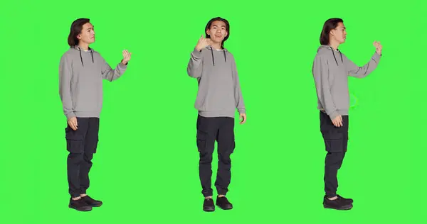 Asian man asking person to come over, encouraging people to get closer while he is standing against full body greenscreen. Adult in casual clothes urging someone to approach him.